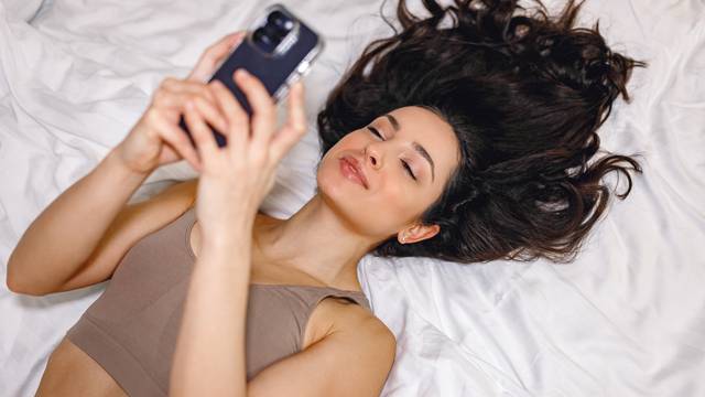 Happy pretty woman holding smartphone while typing. Lady in underwear lying on bed and looking on screen