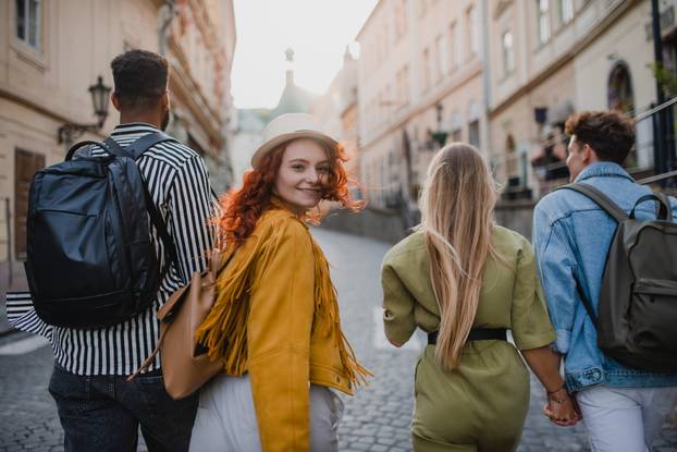 Rear view of group of happy young people outdoors on trip in town, walking and looking back.