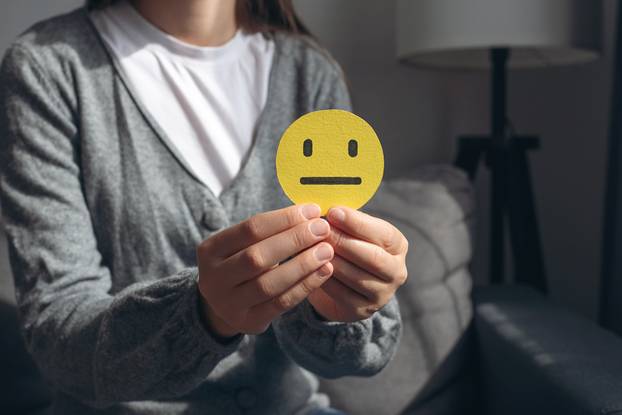Unrecognizable young woman holding small yellow sad face, demonstrate bad emoticon sitting on grey couch. Feedback rating and positive customer review, experience, satisfaction survey concept