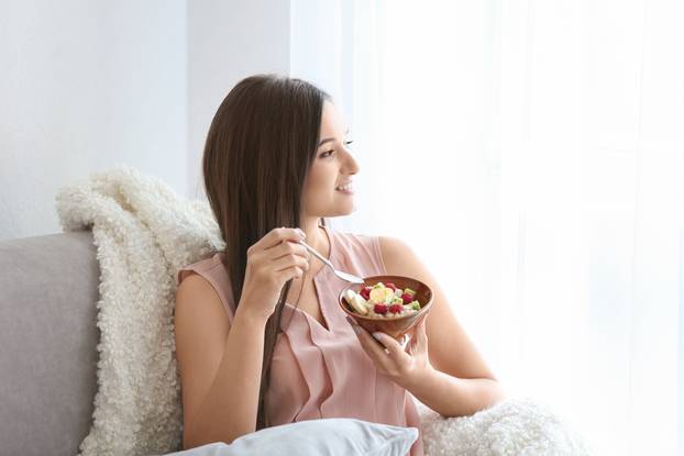Young,Woman,Eating,Oatmeal,On,Sofa,At,Home