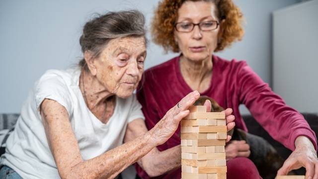 Elder woman and her adult daughter together with dachshund dog spend time together at home playing board game collecting wooden blocks in tower. Jenga game. Theme is dementia and alzheimer's