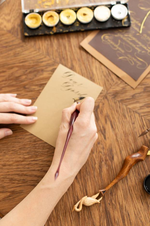 Attractive woman calligrapher is holding a pen with ink and signs a card in handwritten font. Close-up. Soft focus.