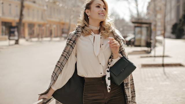 Attractive curly blonde woman in checkered tweed coat, white blouse and brown pants smiles sincerel
