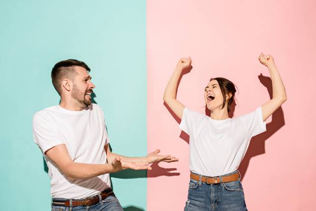 Closeup portrait of young couple, man, woman. One being excited happy smiling, other serious, concerned, unhappy on pink and blue background. Emotion contrasts