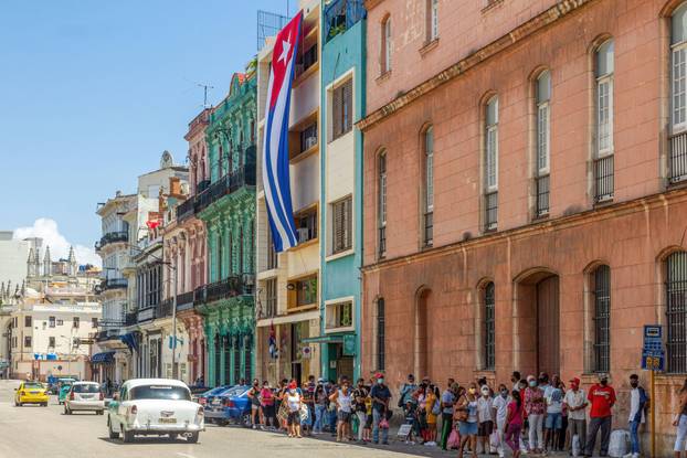 Large group of people waiting for a public omnibus in a bus stop. A large Cuban flag hangs from a government building. An old American car drive in th