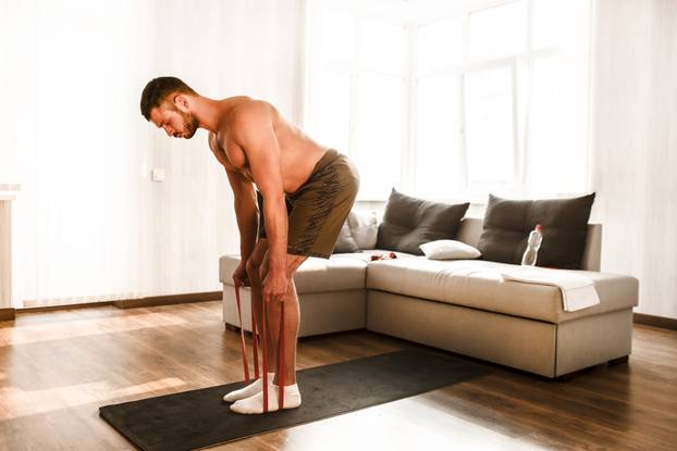 T-shirtless guy sportsman in go in for sport at home. Young man doing sport workout in room during quarantine. Stand on yoga mat and stretch leg on back using resistance band.