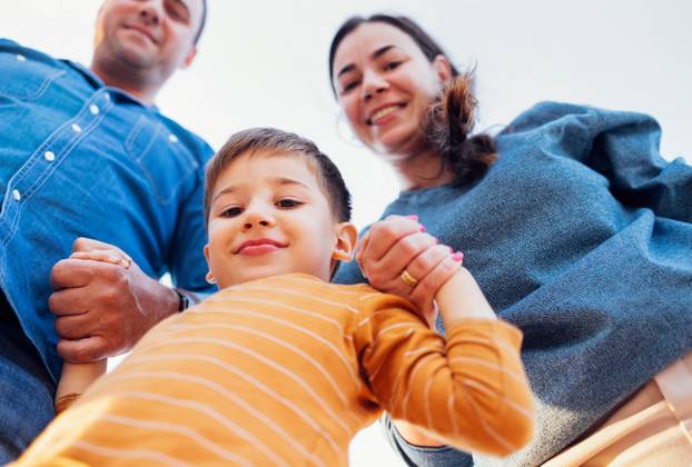 Positive white parents holding hands of their little son. Happy family on a walk. Mom, dad and son in casual clothes look at the camera and smile.