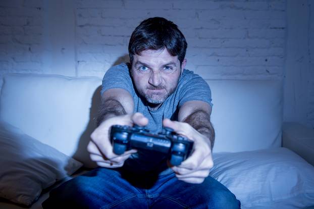 young excited man at home sitting on living room sofa playing video games using remote control joystick