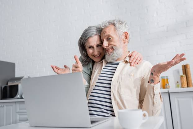 Positive woman hugging confused husband near laptop and cup of coffee in kitchen