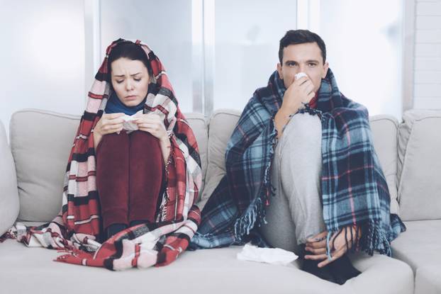 The couple is sitting on the couch wrapped in blankets. Man and woman are sick. The man flies out. Woman holding napkin