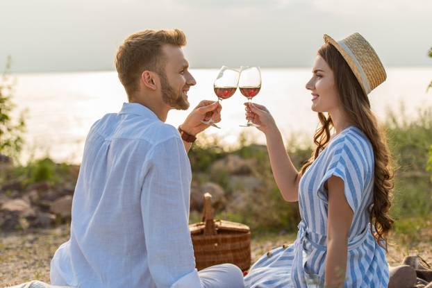 side view of cheerful couple clinking glasses with red wine while sitting near lake