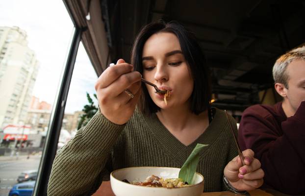 Pretty young woman tastes new meal. Brunette woman eats asian food in a restaurant paying attention to unknown taste while blond man sits near her. Panoramic windows on background.