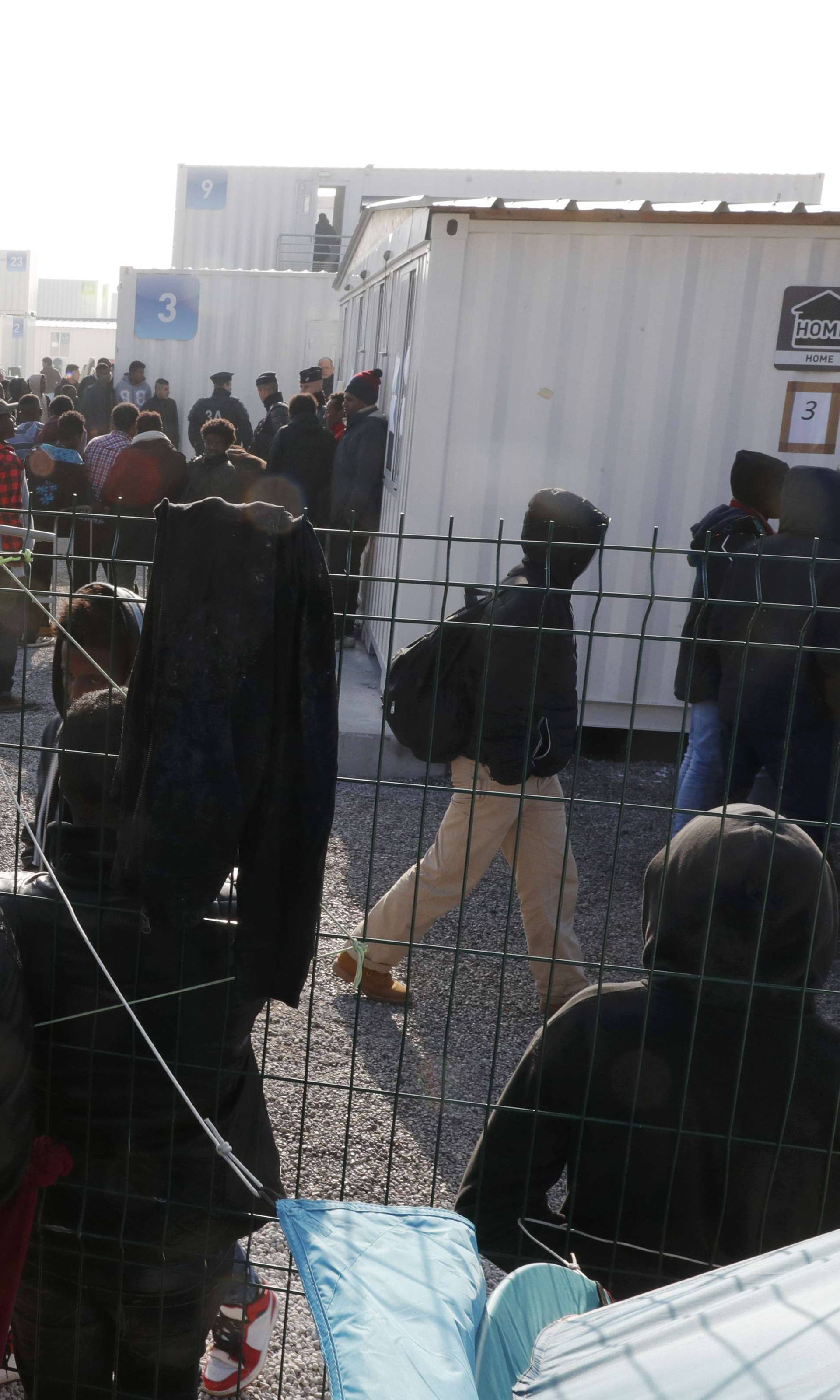 A small group of migrants stand at a fence and look as other migrants gather by the containers used as temporary housing on the eve of the evacuation of the makeshift camp called the "Jungle" in Calais