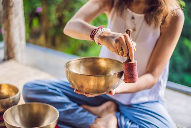 Woman playing on Tibetan singing bowl while sitting on yoga mat against a waterfall. Vintage tonned. Beautiful girl with mala beads meditating