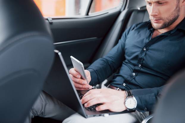 A young businessman working on laptop and talking on the phone while sitting in the car's back. Works in motion, appreciates its time