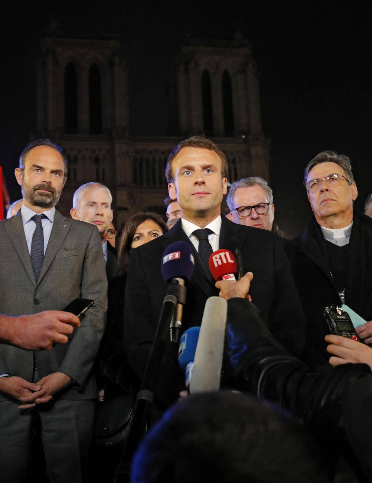 French President Emmanuel Macron speaks as Prime Minister Edouard Philippe and Archbishop of Paris, Michel Aupetit, stand in front of the Notre Dame Cathedral where a fire burns in Paris