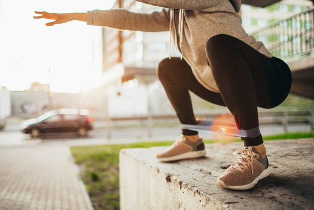 Fit young woman doing squats exercise with fitness gum outdoor in urban environment under the bridge. Slim fit girl stretching outside on a sunset.