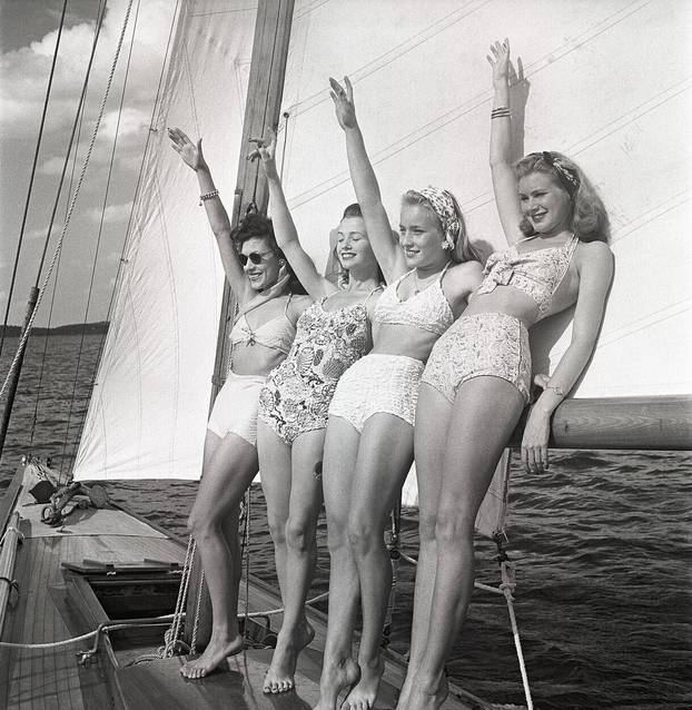 1940s sailing boat. Four young women are standing and leaning against the sails of the boat waiving as you should when you meet another boat. They are wearing the typical 1940s bathingsuit and bikini with high waist. Sweden 1946 Photo Kristoffersson ref A