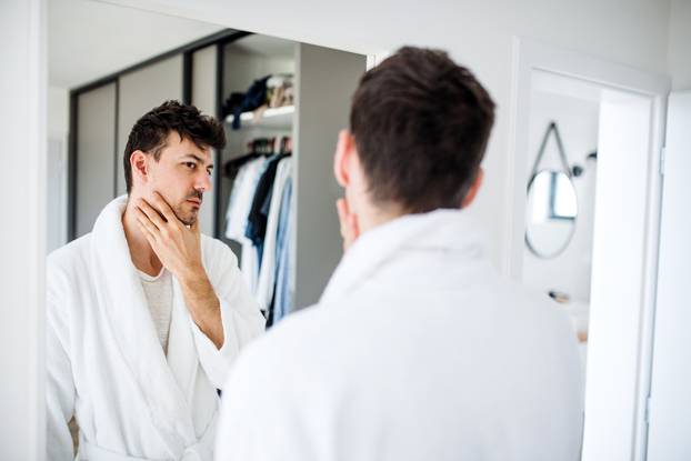 Young man looking in mirror in bedroom in the morning, daily routine.