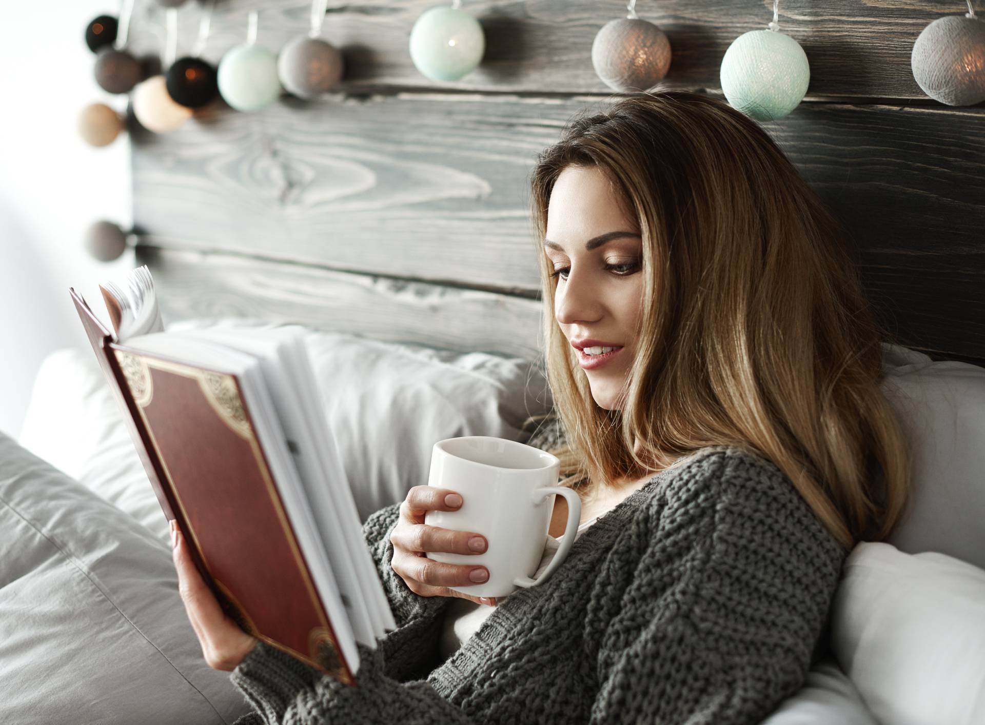 Woman drinking coffee and reading book on bed
