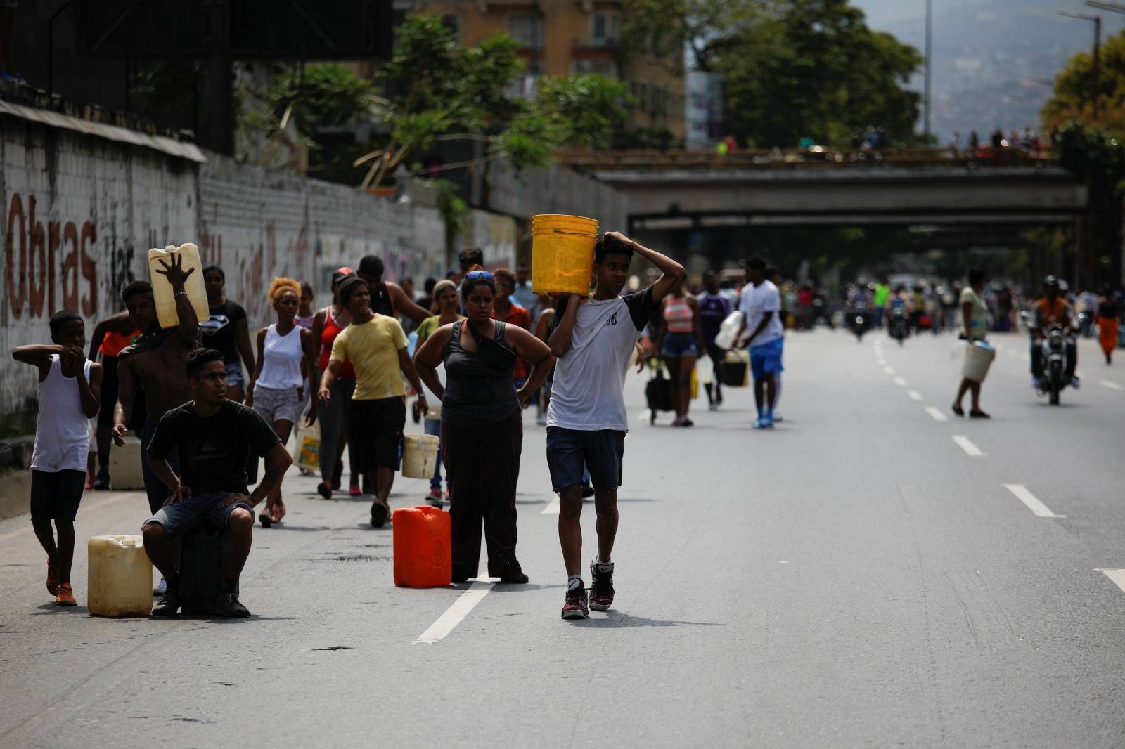 People walk on a highway, carrying containers with water collected from a sewage drain in Caracas