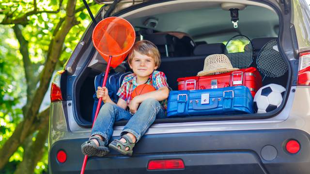 Adorable little kid boy sitting in car trunk just before leaving for summer vacation with his parents. Happy child with suitcases and toys going on journey