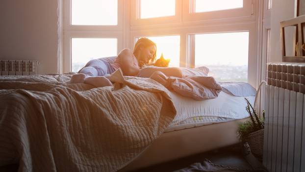 Young pretty happy woman laying in bed with maine Coon cat by the window during sunset with lens flare effects