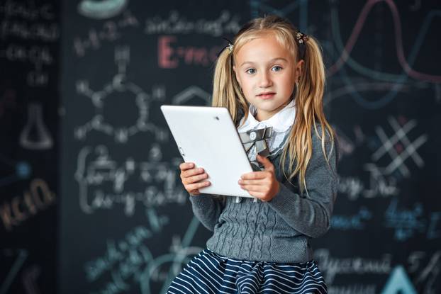 Little blond Girl in school uniform holding white tablet pc in chemistry class. Chalkboard with school formulas background.
