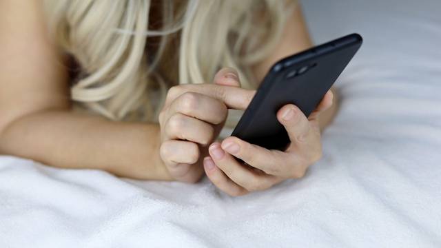 Blonde girl with smartphone laying on the bed. Concept of online addiction, social media, sms messages
