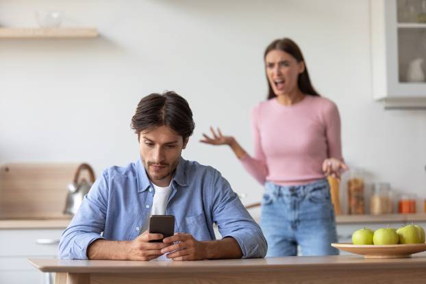 Sad unhappy young caucasian husband with smartphone ignoring upset angry scolding wife