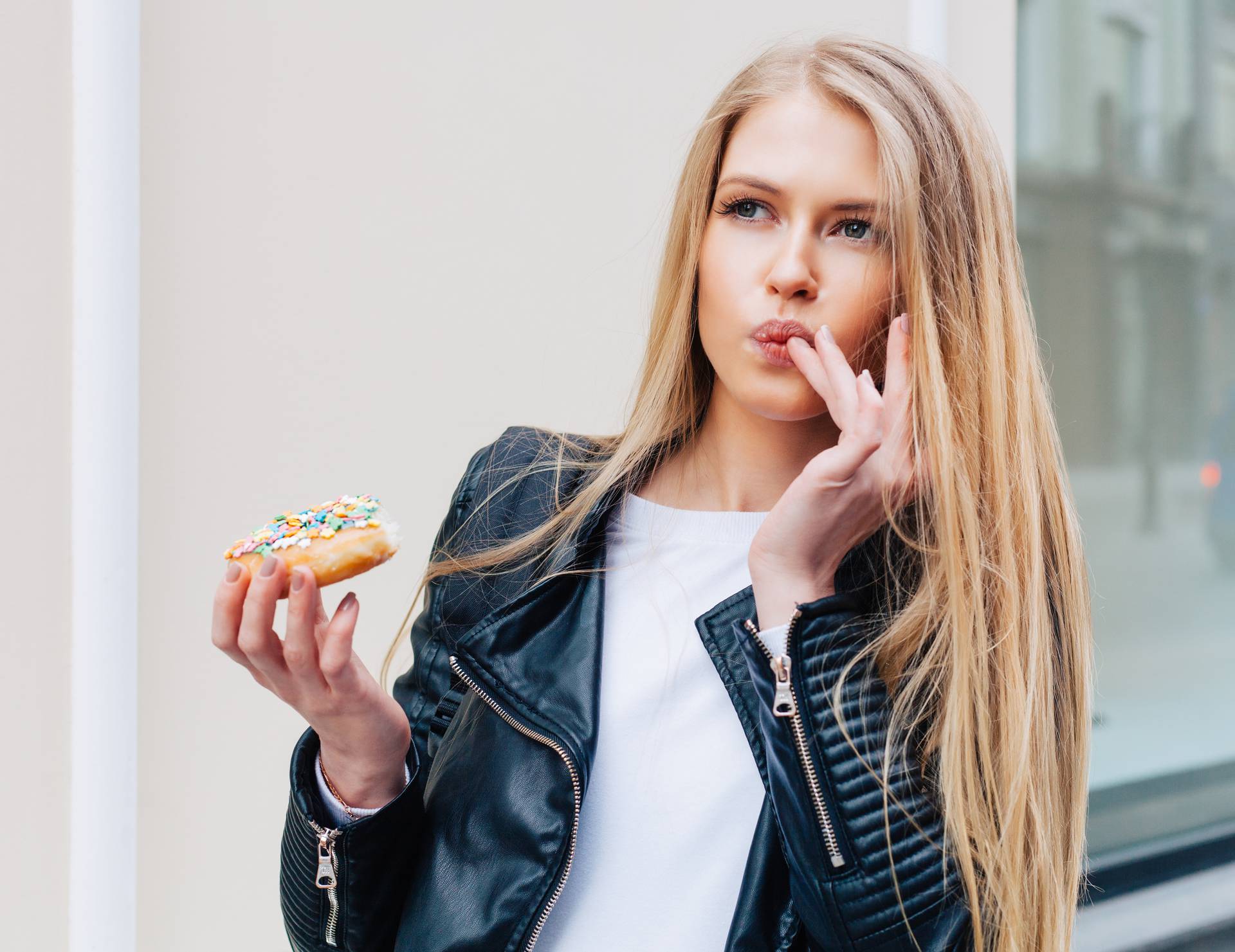 Beautiful young sexy woman eating a donut, licking her fingers taking pleasure a European city street. Outdoor. Warm color.