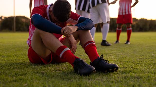 Caucasian sad male athlete sitting on grassy land with team players in background at playground. Rivalry, sunset, frustration, loss, unaltered, soccer, competition and team sport concept.