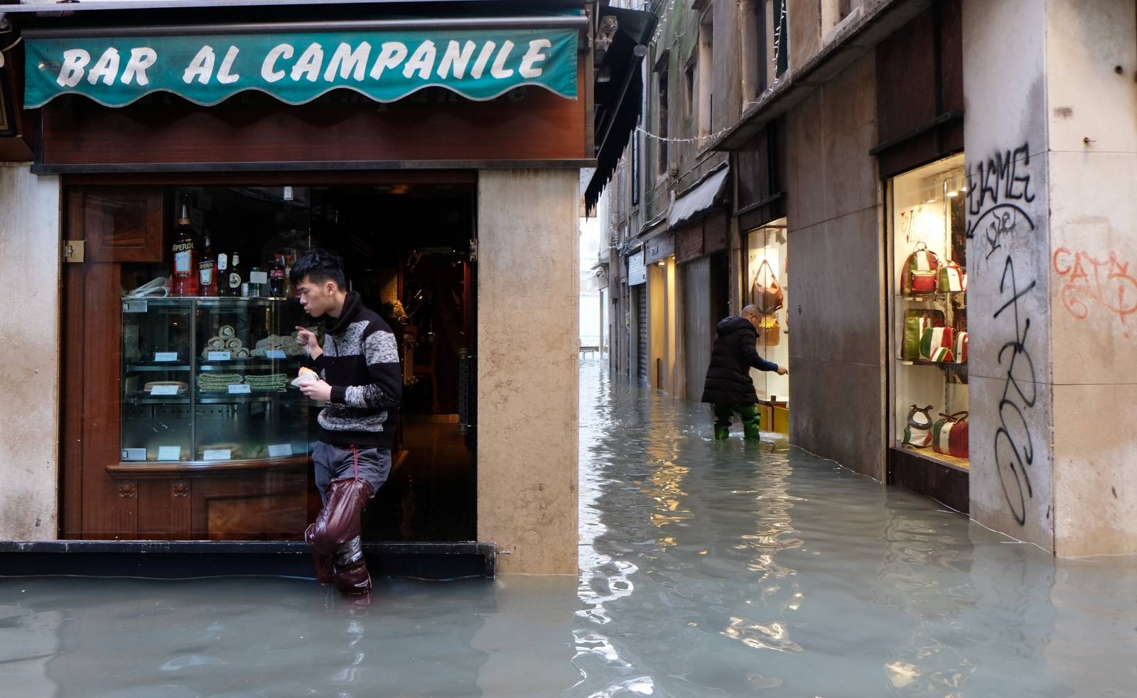 A man wearing rubber boots stands outside a bar on a flooded street during high tide in Venice