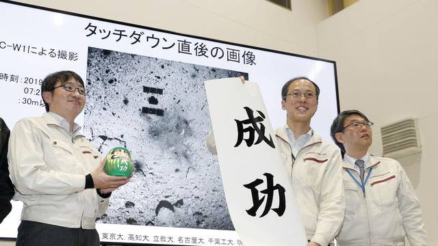 JAXA associate professor Yuichi Tsuda holds a banner reading 'success' in front of an image of the Hayabusa 2 space probe's landing on the Ryugu asteroid in Sagamihara