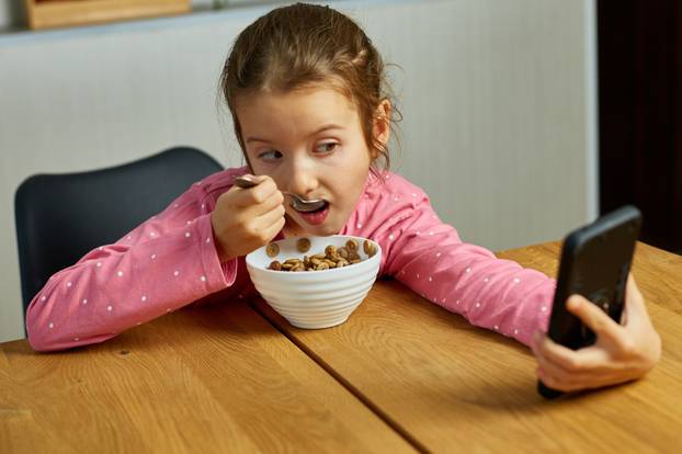 Little girl eating cereal with milk and watching video on smartphone