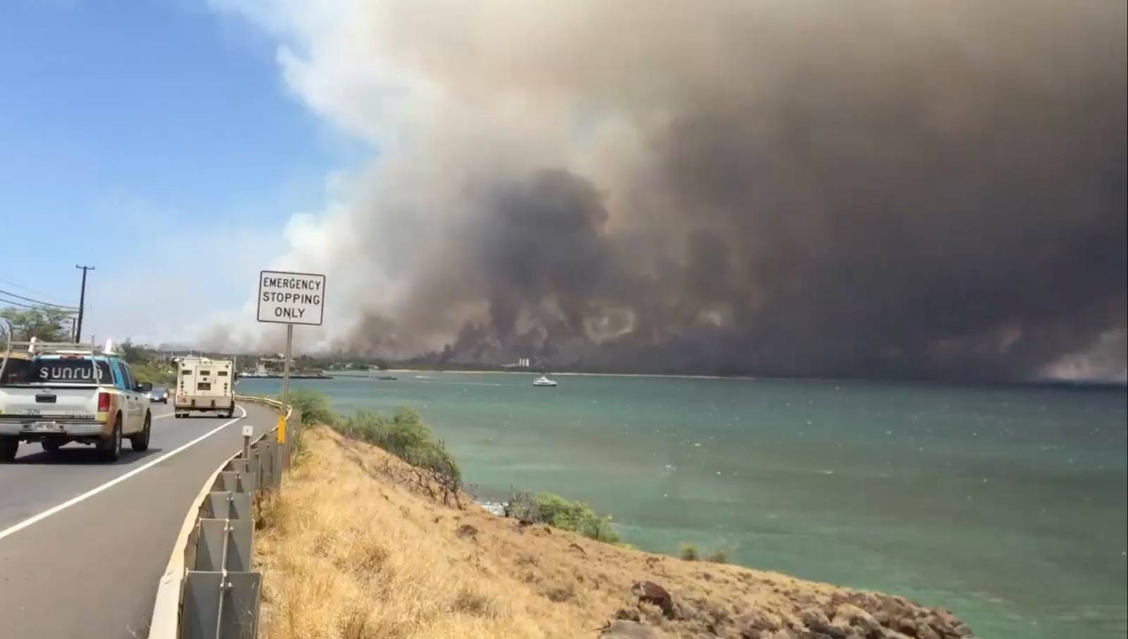 A plume of smoke spreads across the sky during a wildfire in Kihei, as vehicles move along a road in Maui