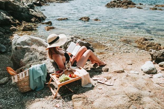 Woman in bikini and hat alone at beach reading electronic book,