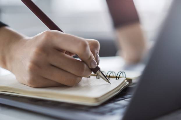 Close-up shot of a woman's hand writing in a notepad on a modern laptop, with a blurred background