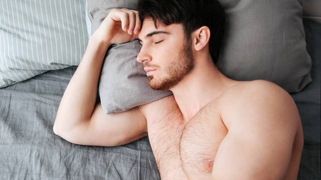 Single young muscular man sleeping in bed. He hold head on pillow. Young man is naked. Lower part of body covered with grey blanket.