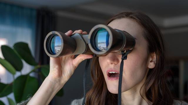 Portrait of a surprised brunette with binoculars looking out the window, spying on neighbors