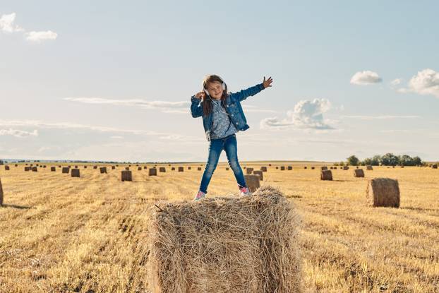 A cute little girl listens to music and dances on a haystack.