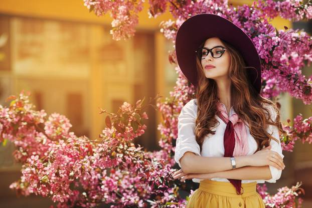 Outdoor portrait of young beautiful fashionable lady posing near flowering tree. Model wearing stylish accessories and clothes. Girl looking aside. Female beauty, fashion concept. City lifestyle. Copy space for text