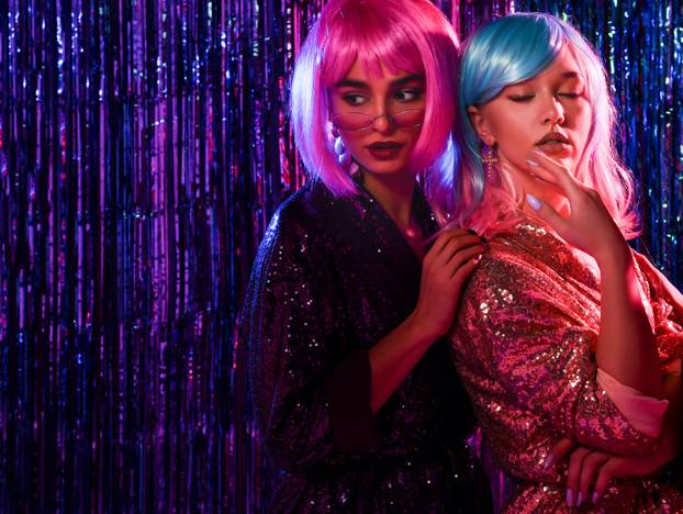 Portrait of happy smiling girls in wigs and stylish glamorous dresses with sequins, in the neon light of a disco. Celebrate, have fun, celebrate something.