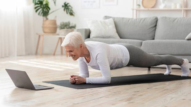 Sporty senior woman doing yoga plank while watching tutorial on laptop