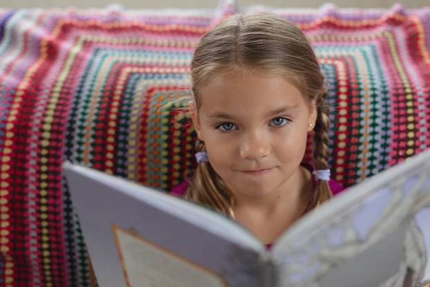Font view of cute Caucasian girl looking at camera while reading a book on sofa in a comfortable home