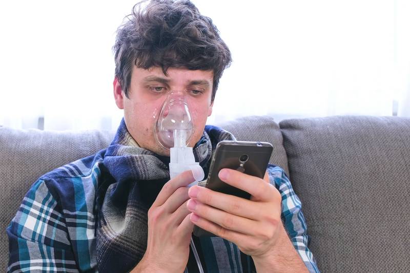 Use nebulizer and inhaler for the treatment. Sick man inhaling through inhaler mask and looking at mobile phone sitting on the sofa.