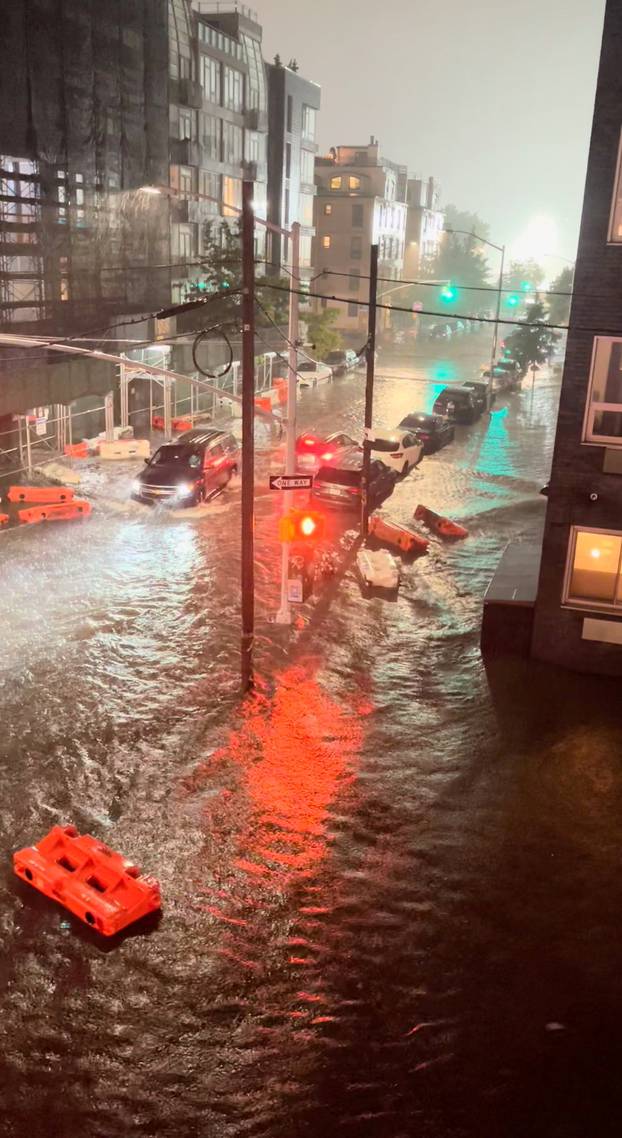 A vehicle moves along a flooded road as safety barriers float in floodwaters in Williamsburg