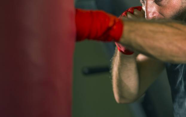 lifestyle gym portrait of young attractive and fierce looking man training boxing at fitness club doing heavy bag punching workout with wrist wraps in badass fighter look 