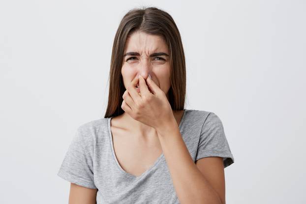 What a smell. Close up of young unhappy caucasian student girl with long hair in casual t-shirt squeezing nose with fingers, looking in camera with disgust expression, feeling bad about bad smell on street.