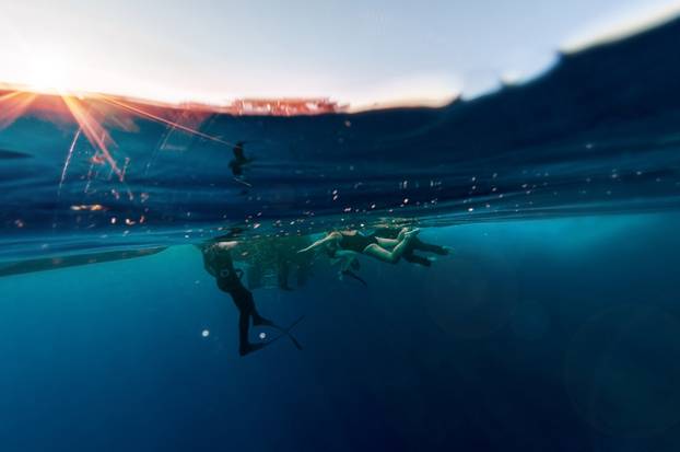 A close view under and above water surface on scuba divers group who climbed after explored deep dark ocean blue water. Underwater fauna and flora, marine life. Active life. Air bubbles.